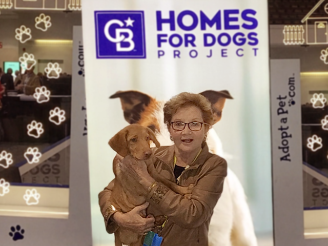 A woman stands holding a tan colored puppy in front of a large sign that features a photograph of a small dog and the words Homes For Dogs Project and adopt a pet dot com